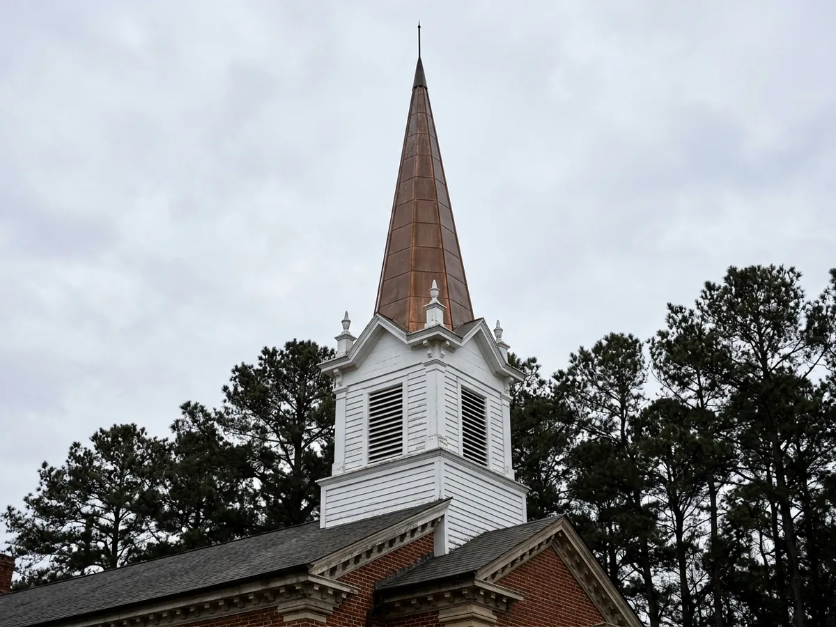 White wooden steeple with a pointed copper-clad spire rising from the ridge of a brick church, flat-seam copper panels on the spire faces