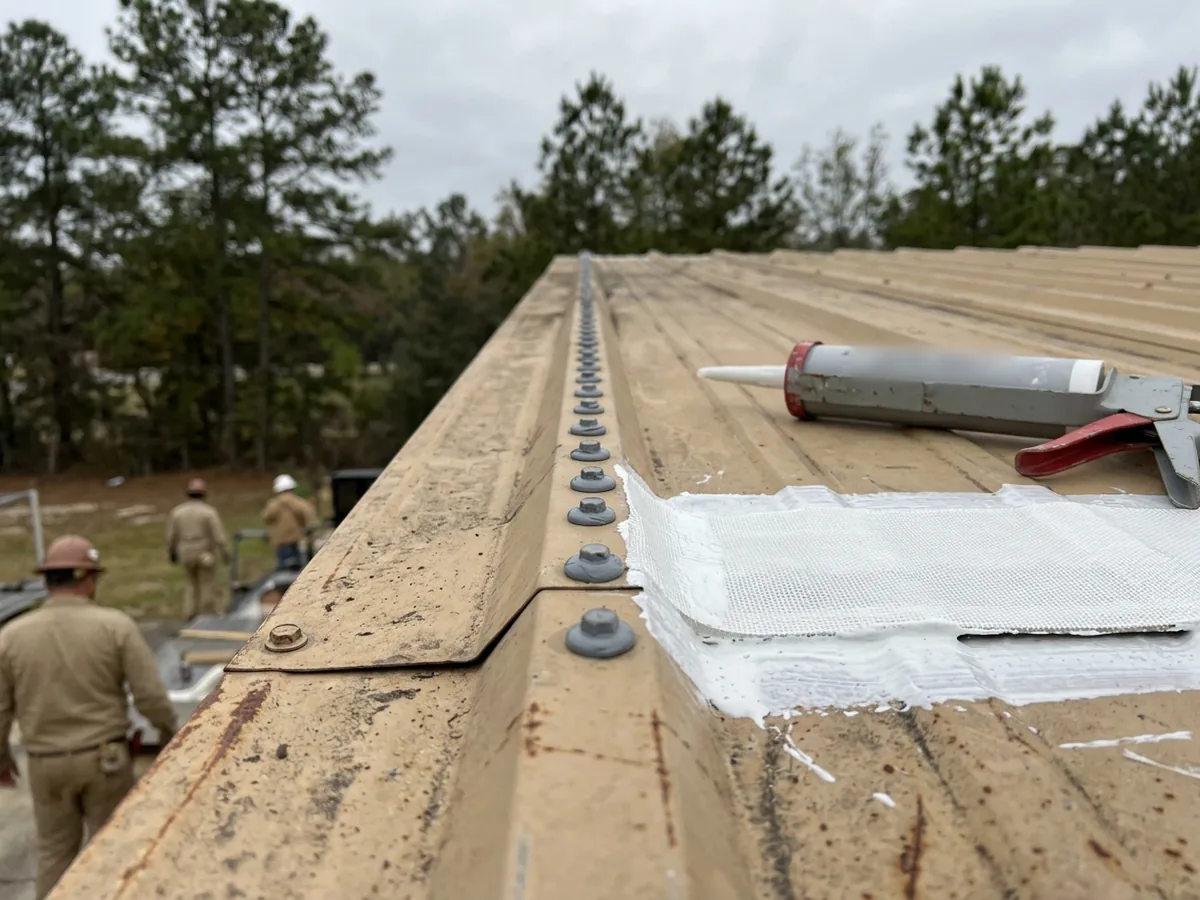 Close view of ribbed metal roof panels with every screw head capped in grey sealant and a strip of white reinforcing fabric bedded along an end lap