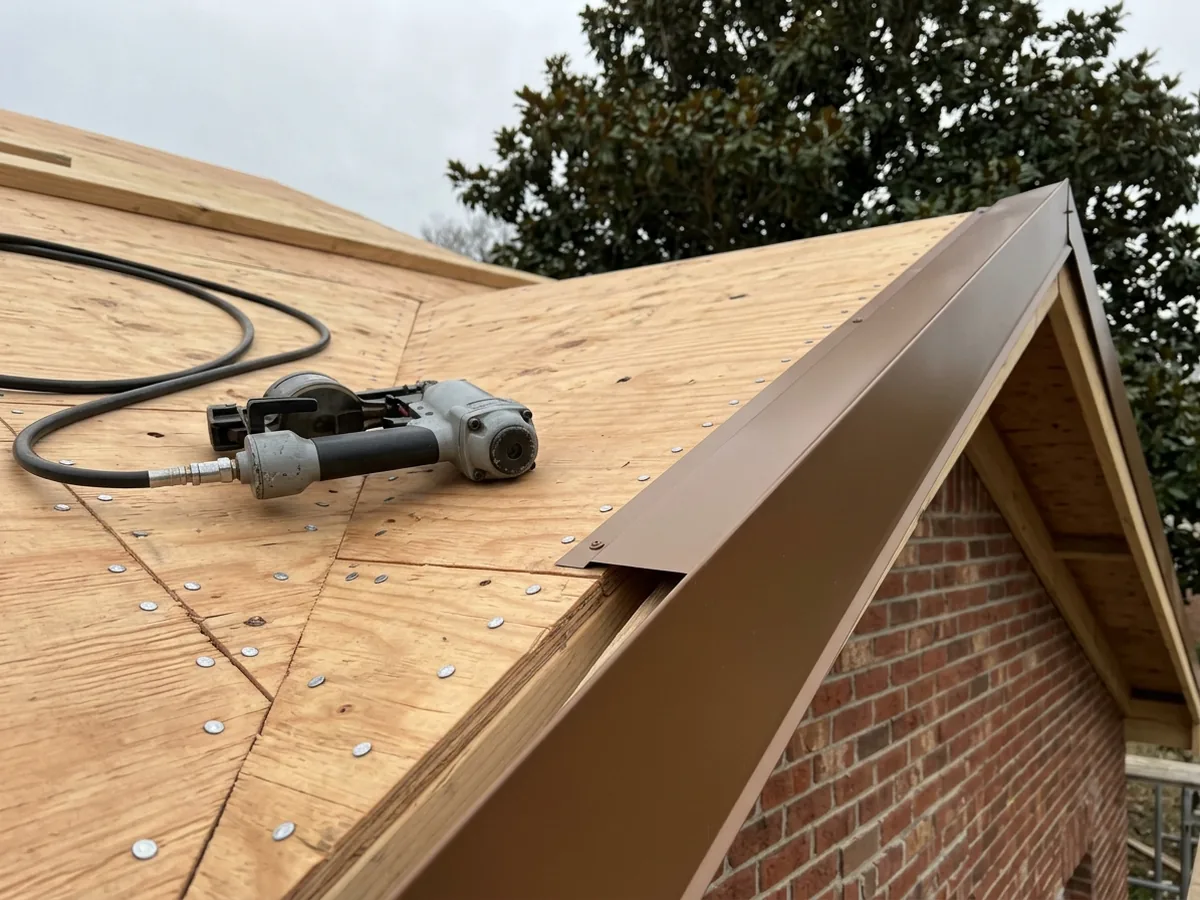 Close view of a roof eave with a wide new metal drip edge and rows of ring-shank nails in fresh plywood decking, an air nailer resting on the deck